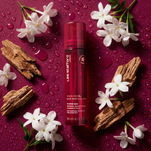 Red bottle of Lee Stafford hair product surrounded by white flowers and wooden elements on a red background