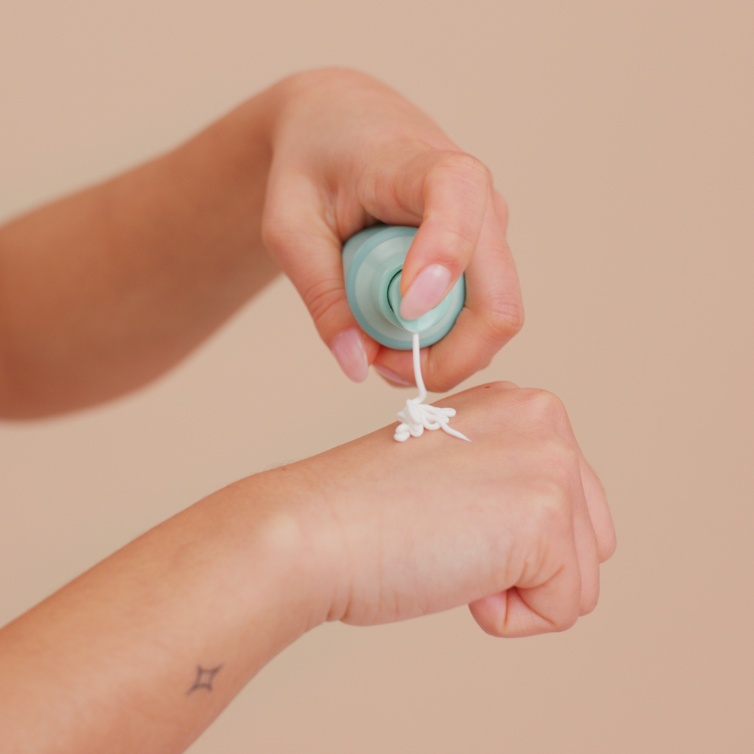 Hand applying a small container of cream to another hand against a beige background
